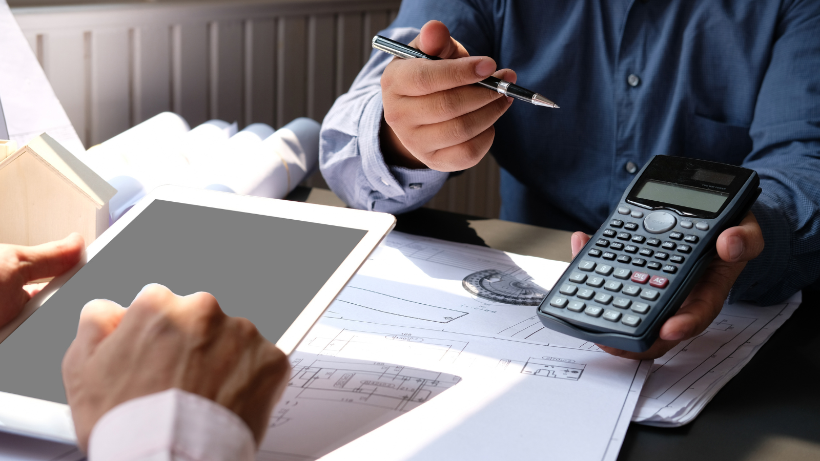 Hands reviewing blueprints with a calculator and tablet